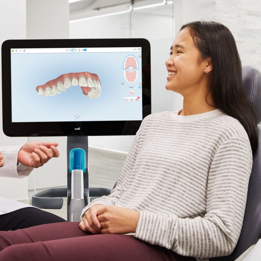 A dentist and a patient review the patient's oral health. A screen shows a digital rendering of her upper teeth.